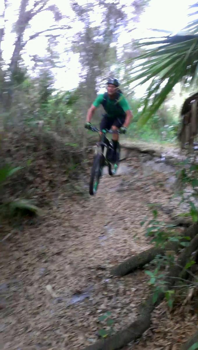 A mountain biker in a green shirt is captured mid-air while jumping over a small obstacle on a dirt trail surrounded by trees and foliage. The image has a blurred effect, emphasizing the action of the jump. North Port Mountain Bike Trails mountain bike trail.
