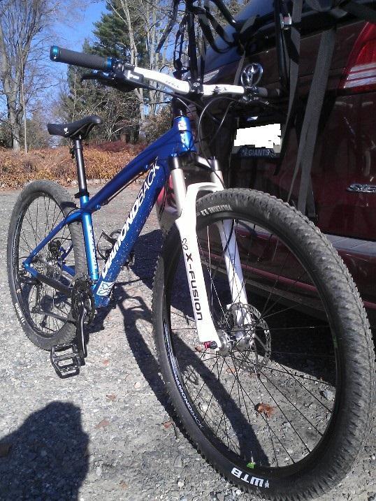 Diamondback Overdrive Sport: A blue Diamondback mountain bike with a white X-Fusion front fork parked on gravel, with trees in the background and part of a vehicle visible to the side.