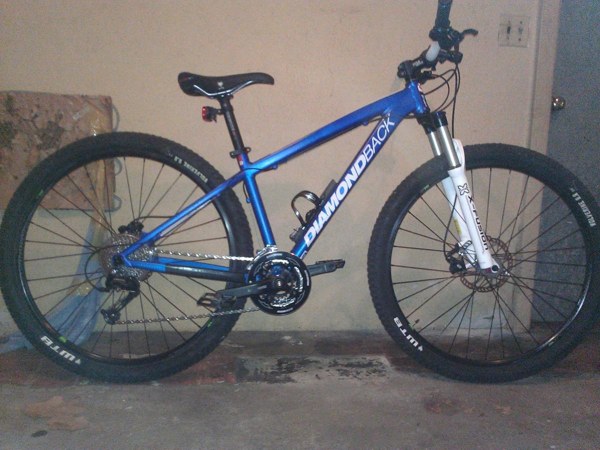 Diamondback Overdrive Sport: A blue Diamondback mountain bike with wide tires, front suspension fork, and a chain drive system is positioned upright against a wall in a garage setting. The background features a partially visible wall and floor.