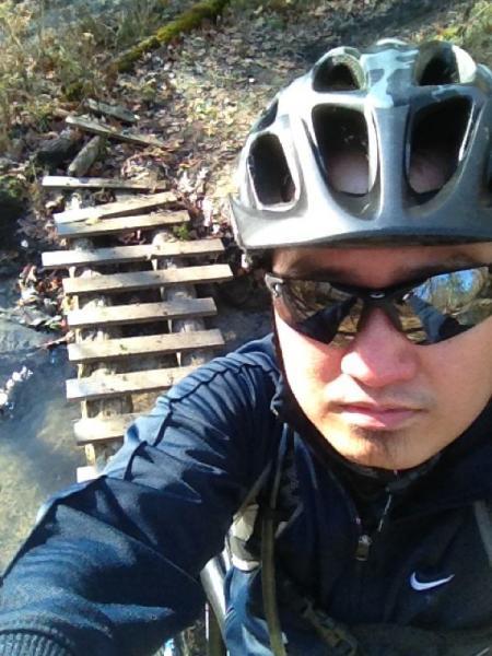 A person wearing a black helmet and sunglasses poses for a selfie while biking on a trail. In the background, there is a narrow wooden bridge made of slats over a stream, surrounded by autumn foliage and a natural setting. Cemetery Loop mountain bike trail.