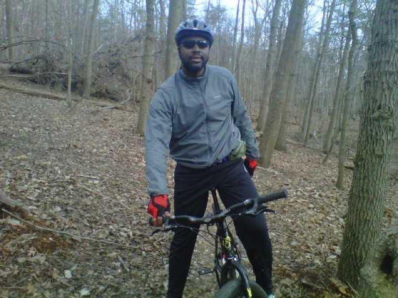 A person wearing a gray jacket, black pants, and a helmet stands next to a mountain bike in a wooded area, surrounded by trees and fallen leaves. The individual is smiling and wearing sunglasses and gloves, suggesting they are enjoying a cycling activity outdoors. Fairland Recreational Park mountain bike trail.