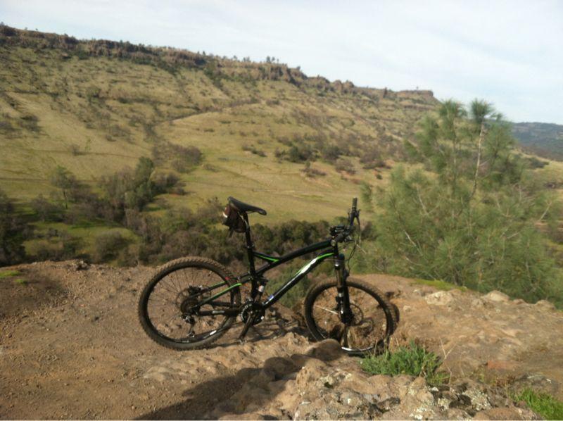 A mountain bike parked on rocky terrain overlooking a scenic landscape of rolling hills and sparse vegetation under a partly cloudy sky. Upper Bidwell Park mountain bike trail.