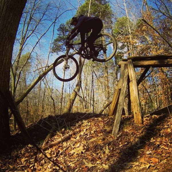 A cyclist in mid-air performing a jump on a mountain bike, soaring over a wooden ramp in a forested area with fallen leaves covering the ground. Trees in the background are bare, suggesting a late autumn setting with a clear blue sky above. Salem Lake mountain bike trail.
