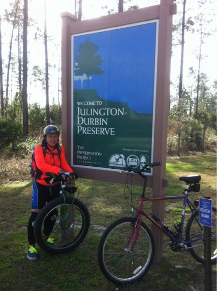 A person in active wear stands next to two bicycles in front of a large sign that reads "Welcome to Julington-Durbin Preserve." The sign features blue and green colors and includes logos related to conservation. The background consists of trees and natural scenery. Julington-Durbin Preserve mountain bike trail.