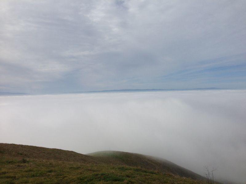 A panoramic view of a cloudy landscape, with a thick layer of fog covering the horizon. Rolling green hills are partially visible in the foreground, while the sky is overcast with gray and blue hues. Alum Rock County Park mountain bike trail.