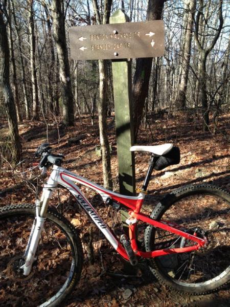 A mountain bike leaning against a trail sign in a forested area. The sign indicates directions for two trails: "Bennett mountain trail" and "Stanley gap trail." The surrounding terrain is covered in brown leaves, and the trees are bare, suggesting it's early spring or late fall. Stanley Gap mountain bike trail.