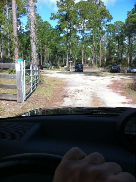 A view of a dirt road leading into a wooded area, taken from the driver's perspective inside a car. On the left, there is a wooden gate, and in the background, several parked vehicles are visible among tall pine trees under a clear blue sky. Moses Creek mountain bike trail.