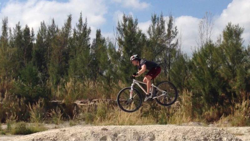 A cyclist performing a jump on a mountain bike, mid-air above a dirt ramp, with lush greenery in the background and a clear blue sky. Virginia Key North Point mountain bike trail.