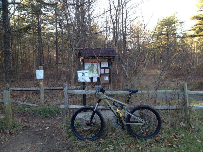 A mountain bike leaned against a wooden fence at the entrance of a forest trail, with a trail map and informational signs visible in the background. The scene is set in a wooded area with bare trees and autumn foliage on the ground. Cemetery Loop mountain bike trail.