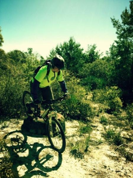 A mountain biker in a bright green shirt and helmet navigates a dirt trail surrounded by vegetation, poised to jump off a small mound. Sunlight filters through the trees, creating a vibrant outdoor scene. Jonathan Dickinson State Park mountain bike trail.