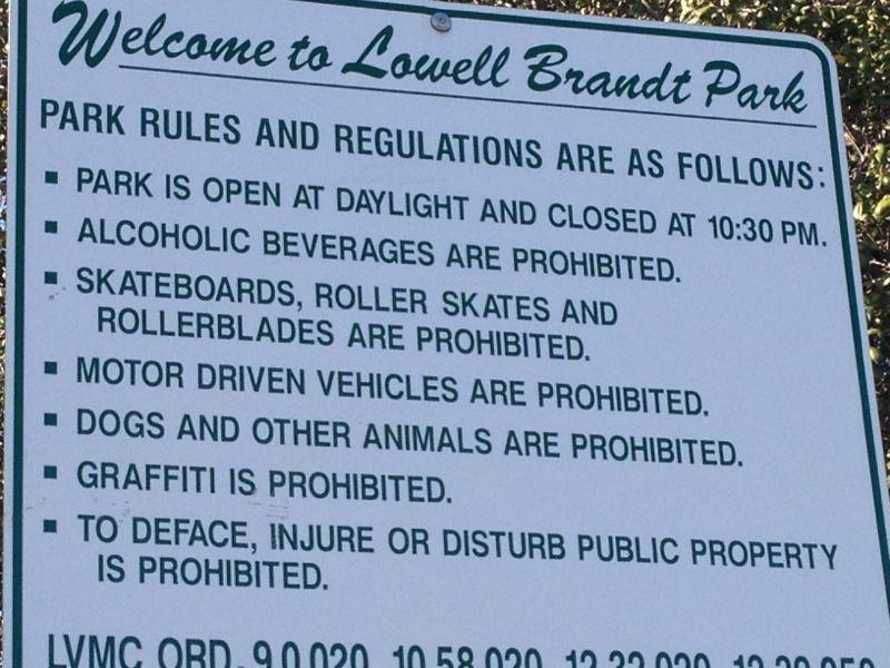 A sign at Lowell Brandt Park displaying park rules and regulations, including hours of operation, prohibited activities such as alcohol consumption, skateboards, roller skates, motor vehicles, pets, and graffiti. Marshall Canyon Park mountain bike trail.