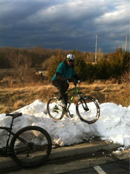 A cyclist in a teal shirt and helmet performs a maneuver on a snowy mound while riding a green mountain bike. In the foreground, another bike is partially visible, and the background features a cloudy sky and a grassy area. Fairland Recreational Park mountain bike trail.