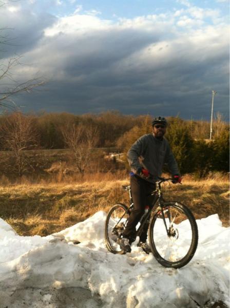 A person wearing a gray sweater and gloves stands on a mound of snow next to their mountain bike, with a backdrop of dark, cloudy skies and a grassy landscape. Fairland Recreational Park mountain bike trail.