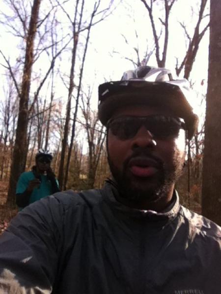 A man wearing a bicycle helmet and sunglasses, standing in a wooded area, with bare trees in the background. Another person is partially visible in the background, also wearing a helmet and a blue shirt, appearing to take a photo. The scene is well-lit, indicating a sunny day. Fairland Recreational Park mountain bike trail.