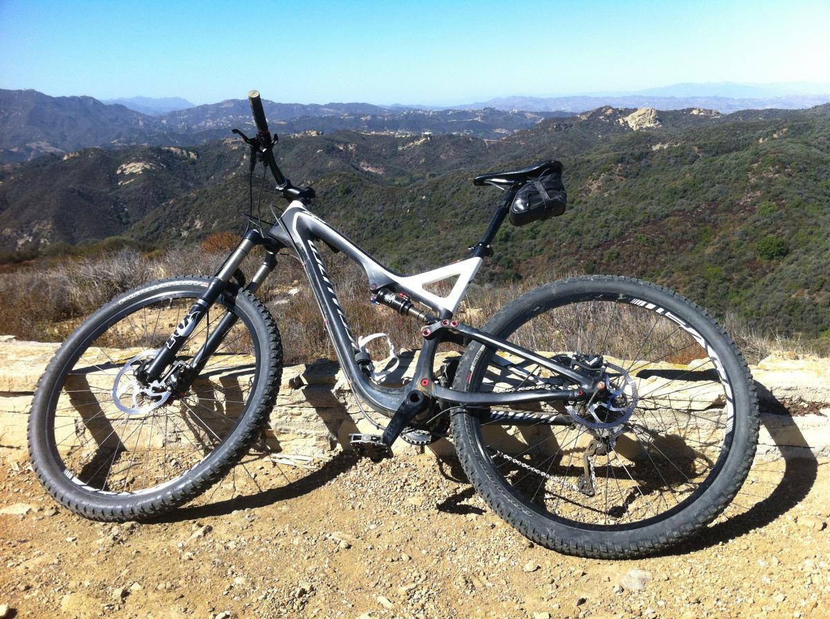 Specialized Stumpjumper FSR: A mountain bike resting on a rocky ledge with a scenic view of rolling hills and valleys in the background. The sky is clear and blue, enhancing the outdoor adventure atmosphere.