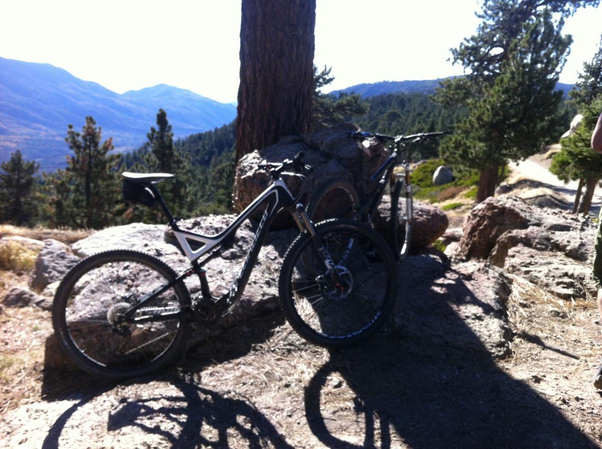 Specialized Stumpjumper FSR: A pair of mountain bikes leaning against a large rock in a scenic outdoor location, surrounded by trees and distant mountains under a clear blue sky.