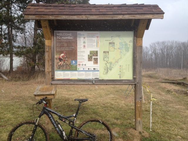 A wooden information board displaying a trail map and guidelines for mountain biking at the location, with a mountain bike leaning against it. The background features a grassy area and trees, indicating an outdoor setting. MoMBA @ Huffman MetroPark mountain bike trail.