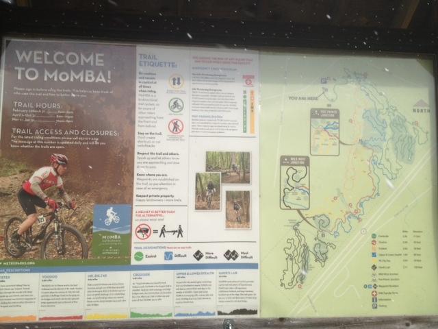A trail information sign for MOMBA featuring trail hours, access details, and etiquette guidelines. The sign includes a map indicating the visitor's location, and images of mountain bikers in a natural setting, emphasizing safety and responsible trail use. MoMBA @ Huffman MetroPark mountain bike trail.