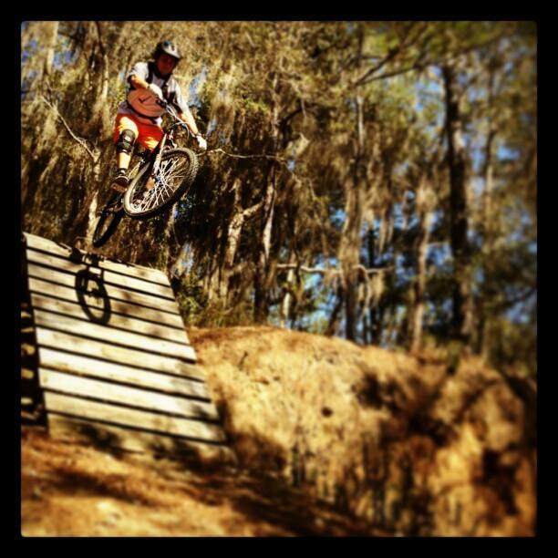 A person wearing a helmet and protective gear is mid-air on a mountain bike, jumping off a wooden ramp in a wooded area. Sunlight filtering through the trees enhances the outdoor setting. Santos mountain bike trail.