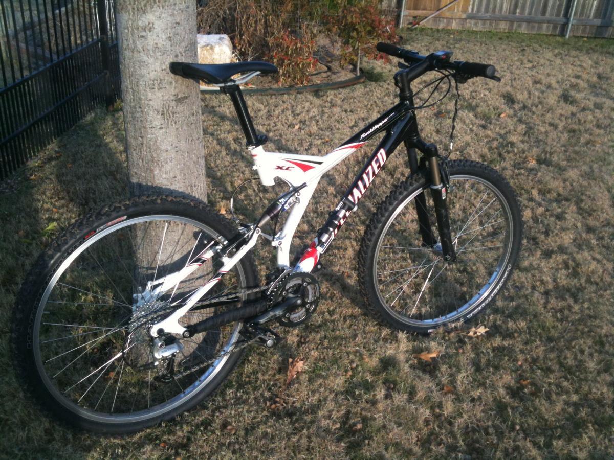Specialized Rockhopper: A mountain bike with a black and white frame, featuring red accents, is leaning against a tree in a grassy yard. The bike has thick tires designed for off-road terrain and a visible gear mechanism. The background includes a black fence and some dried leaves on the ground.