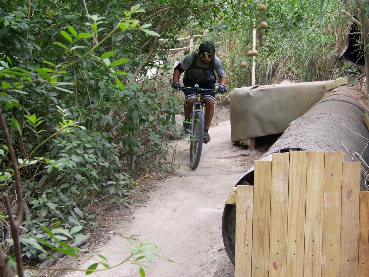 A mountain biker navigating a narrow dirt path surrounded by dense greenery and foliage, with a wooden barrier and a large pipe in the background. Virginia Key North Point mountain bike trail.