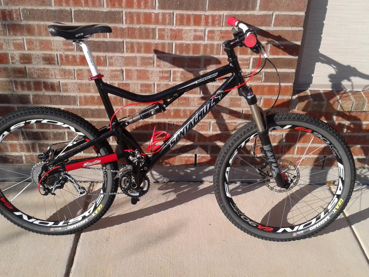 Santa Cruz Superlight: A black and red mountain bike with prominent features, including thick, treaded tires and a suspension fork, positioned against a brick wall. The bike has a sleek design, visible gears, and quick-release wheels, showcasing its readiness for off-road riding.