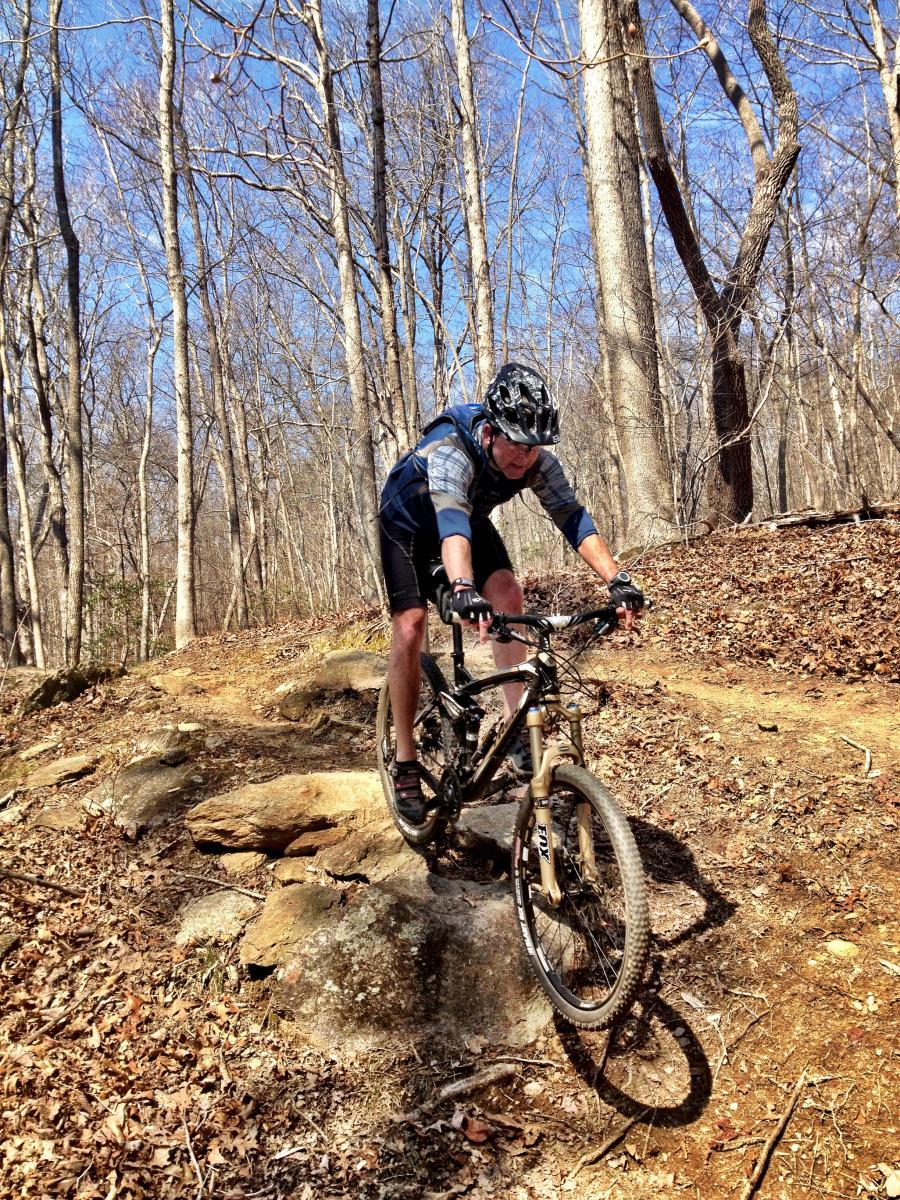 A cyclist navigating a rocky trail in a forest during daytime, surrounded by bare trees and blue sky. The rider is wearing a helmet and cycling gear, focused on maintaining balance as they maneuver over uneven terrain. Chicopee Woods mountain bike trail.