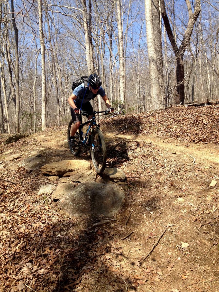 A person riding a mountain bike on a rocky trail in a forest. The cyclist is leaning forward and navigating over a large rock, surrounded by bare trees and a clear blue sky. The ground is covered with leaves and dirt, indicating a natural outdoor setting. Chicopee Woods mountain bike trail.