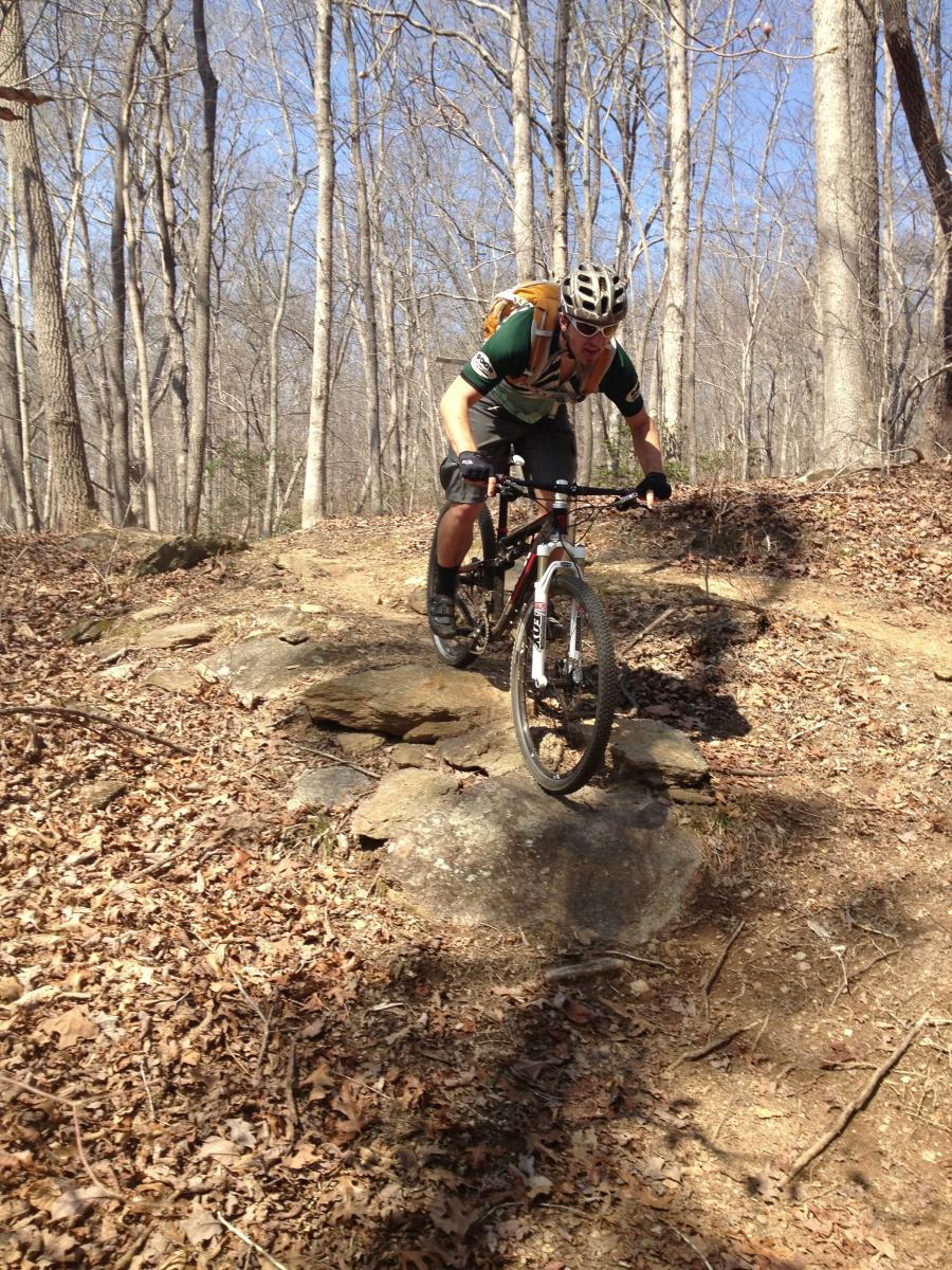 A mountain biker navigating a rocky trail in a wooded area, leaning forward on their bike while balancing on a large stone. The surrounding trees are bare, indicating early spring or late fall, with scattered leaves on the ground. Bright sunlight filters through the branches. The cyclist wears a helmet and a backpack, dressed in shorts and a short-sleeved shirt. Chicopee Woods mountain bike trail.