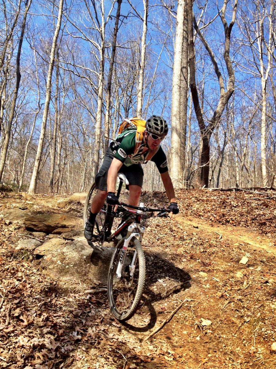 A cyclist navigating a rocky trail in a wooded area on a mountain bike, wearing a helmet and a backpack. The background features bare trees against a clear blue sky, with leaves scattered on the ground. Chicopee Woods mountain bike trail.