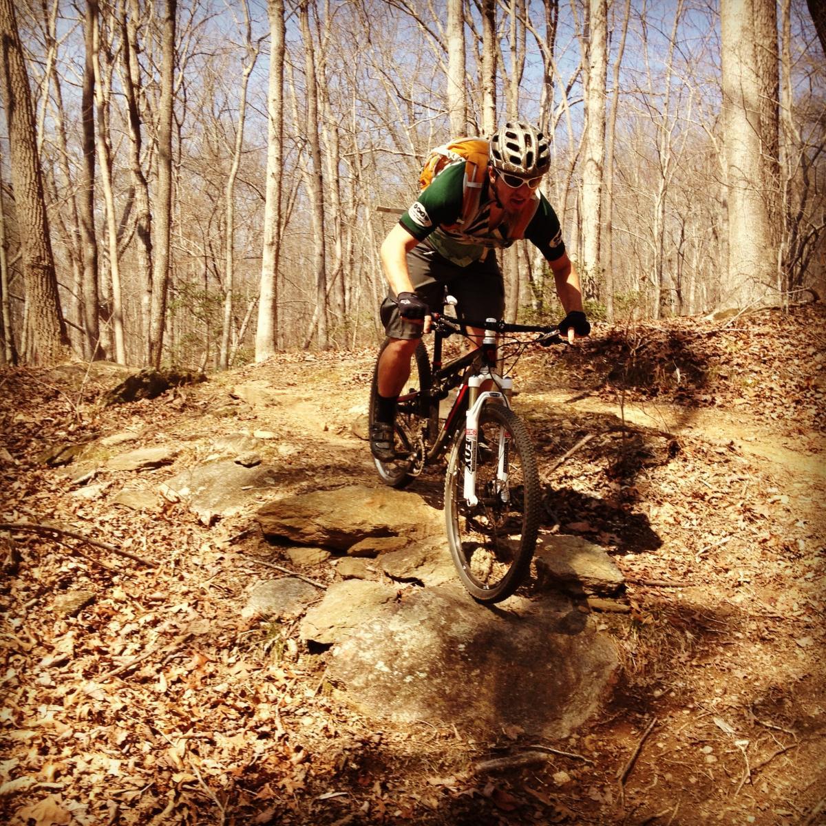A mountain biker navigating over rocky terrain in a wooded area during the daytime. The cyclist is wearing a helmet and protective gear, and the ground is covered in fallen leaves. Chicopee Woods mountain bike trail.