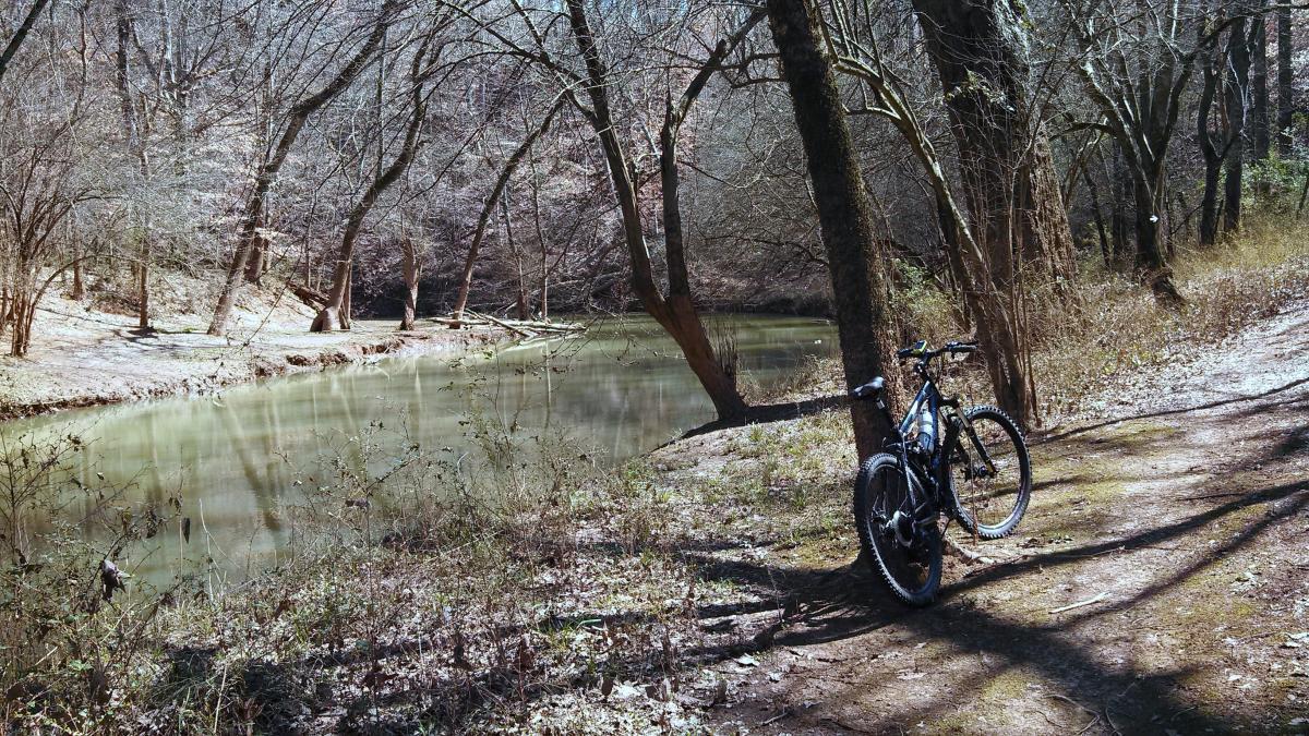 A mountain bike parked beside a calm, greenish river, surrounded by bare trees and a peaceful natural landscape. The scene captures a quiet moment in a wooded area, with sunlight filtering through the branches. Water Tank Trail mountain bike trail.
