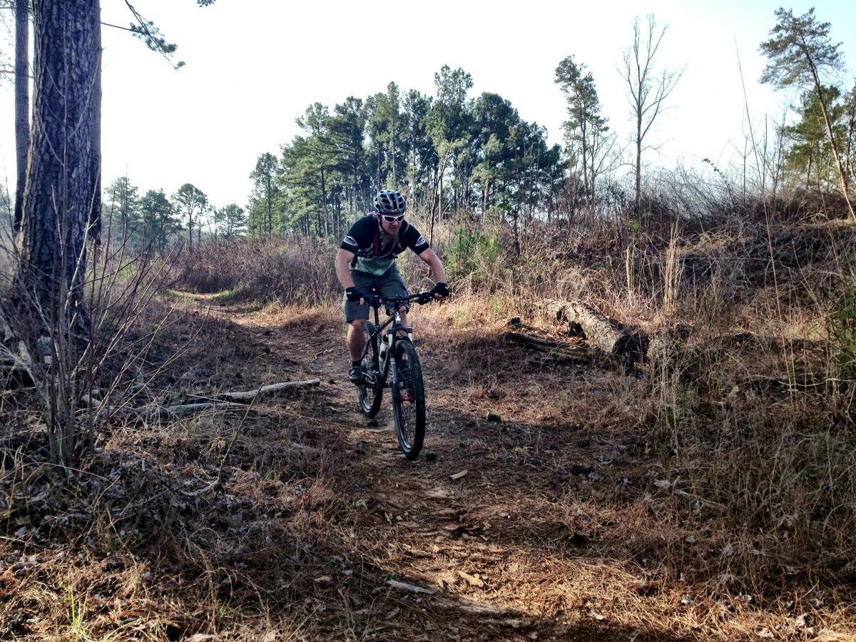 A mountain biker riding on a dirt trail surrounded by trees and underbrush in a forested area. The cyclist is wearing a helmet and sunglasses, focused on navigating the terrain. The scene is sunny and showcases a natural outdoor setting. Lake Russell Loop mountain bike trail.