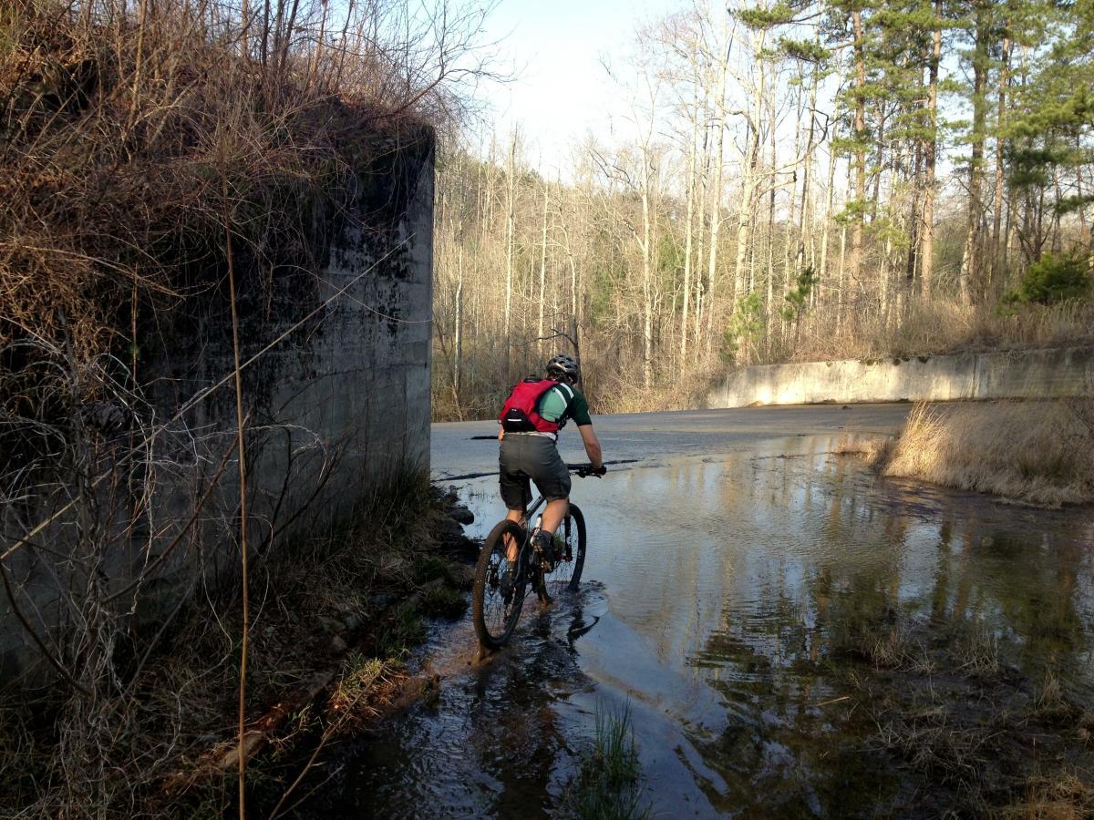 A mountain biker navigating through shallow water on a trail, with a concrete structure to the left and trees in the background. The scene is set in a wooded area during daylight. Lake Russell Loop mountain bike trail.