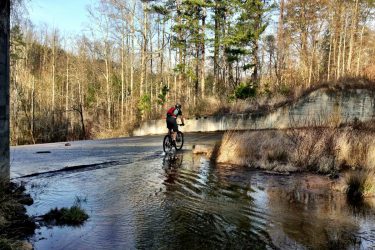 A cyclist riding a mountain bike through a shallow water-covered path in a forested area. The scene features bare trees and a clear blue sky, with a mix of greenery along the sides of the path. Lake Russell Loop mountain bike trail.