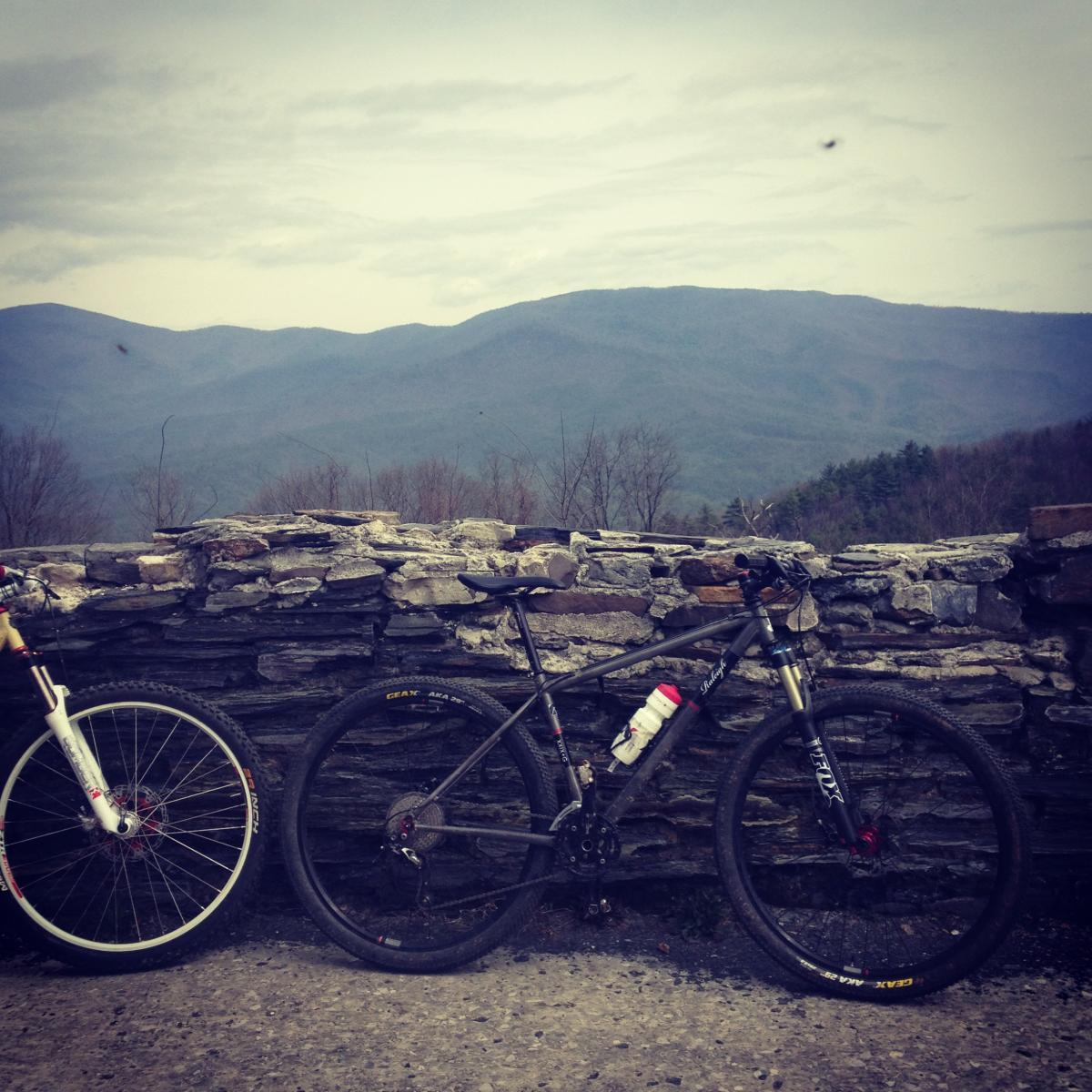 Two mountain bikes are parked next to a stone wall, overlooking a scenic view of rolling mountains and cloudy sky. The foreground features a white bike on the left and a black bike on the right, each displaying distinct designs and a water bottle attached. The surrounding landscape is lush with trees and peaks in the background. Pinhoti Trail mountain bike trail.