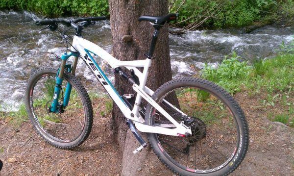 Yeti AS-R (Alloy): A mountain bike resting against a tree near a flowing stream, surrounded by green grass and foliage. The bike features a white frame with turquoise accents and rugged tires suited for off-road terrain.