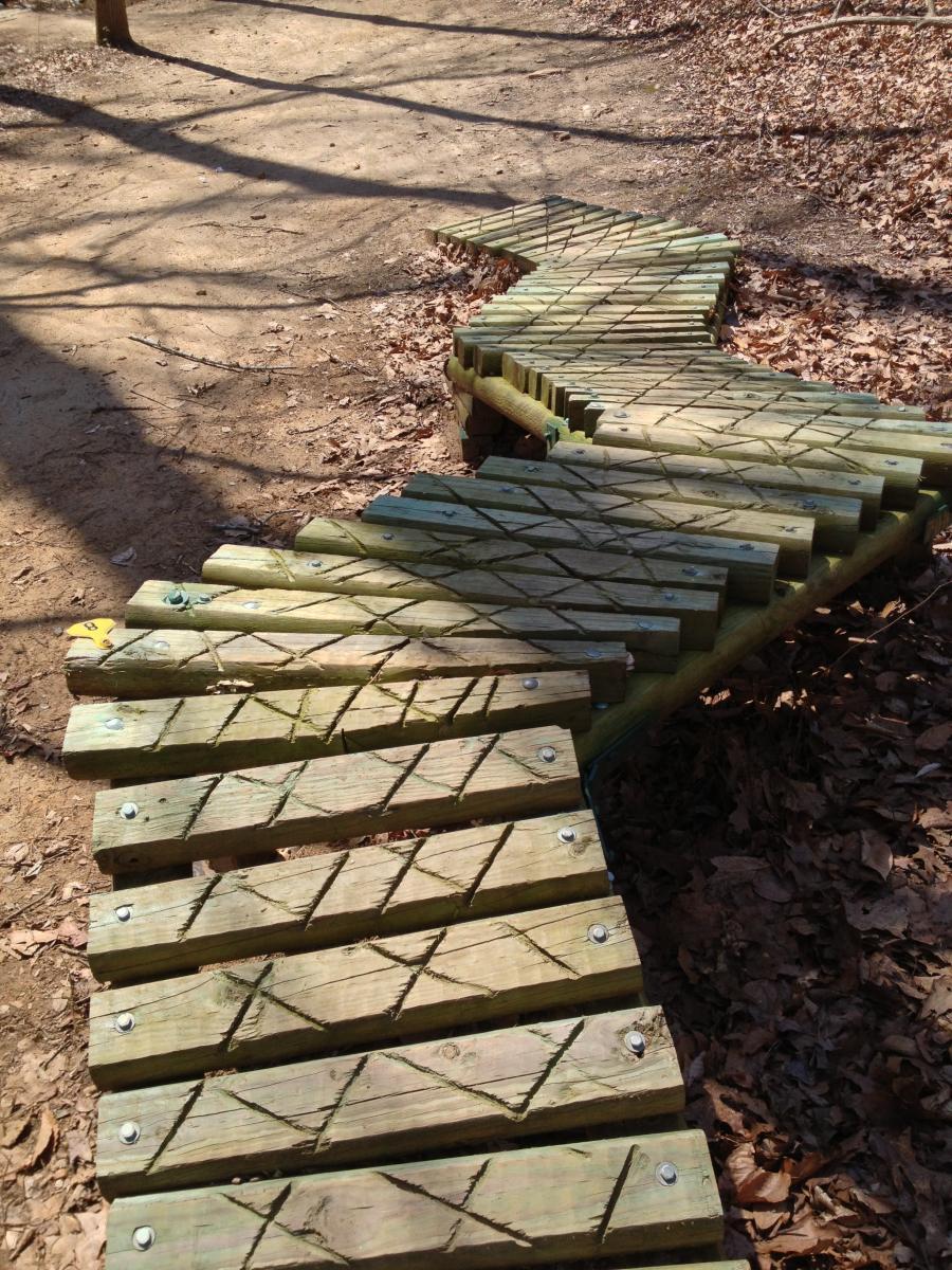 A winding wooden boardwalk made of planks, set against a backdrop of dirt and fallen leaves, leading through a forested area. The path is designed with a crisscross pattern for traction and is surrounded by bare trees, suggesting an early spring season. Angler's Ridge mountain bike trail.