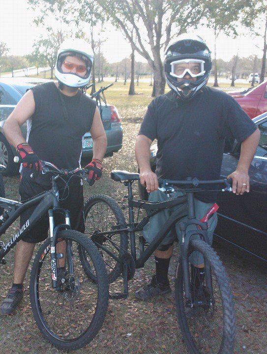 Two men wearing helmets and protective gear stand next to their mountain bikes in a grassy area. The man on the left is wearing a sleeveless shirt and red gloves, and the man on the right is in a black shirt with dark shorts. There are trees and cars visible in the background, indicating an outdoor biking location. Markham Park mountain bike trail.