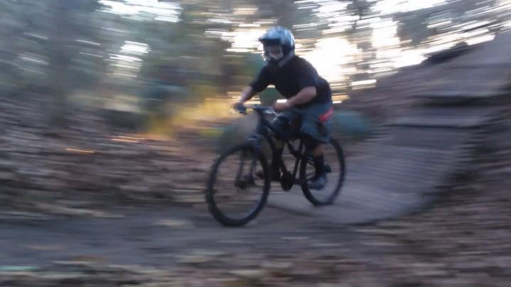 A person wearing a helmet riding a mountain bike on a dirt trail surrounded by trees, captured in motion with a blurred background to emphasize speed. Markham Park mountain bike trail.