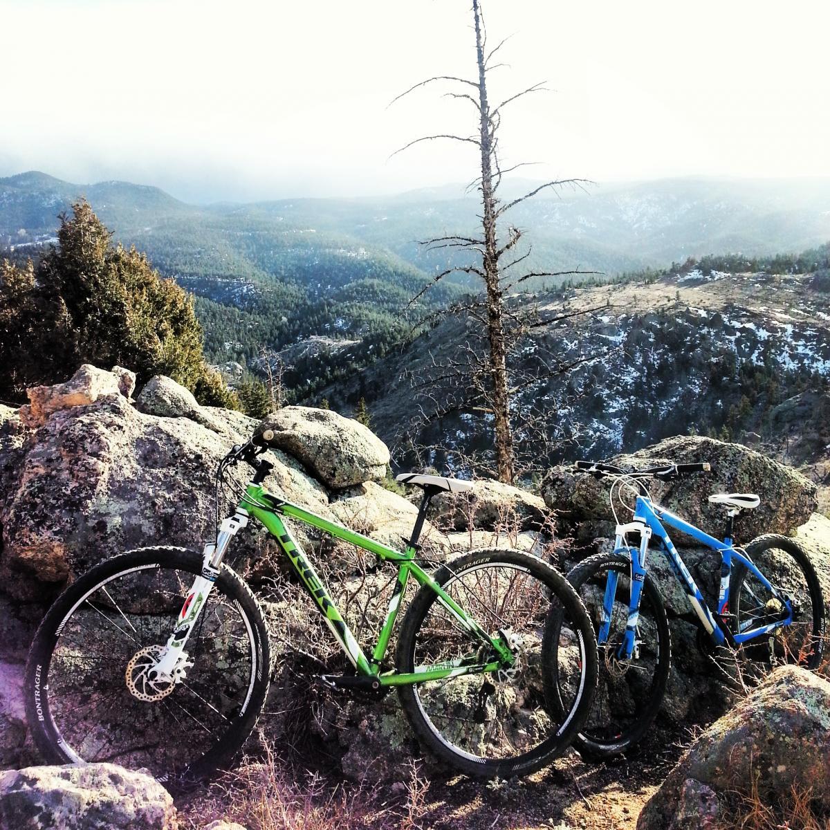 Two mountain bikes, one green and one blue, resting against large rocky outcrops, with a scenic view of rolling hills and pine-covered mountains in the background. A few patches of snow are visible on the terrain under a bright sky. Walker Ranch mountain bike trail.