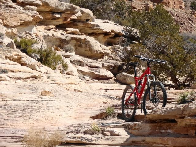 A red mountain bike is parked on rocky terrain, surrounded by textured stone formations and sparse vegetation in a natural outdoor setting. The scene captures the rugged beauty of the landscape, highlighting the bike against the backdrop of cliffs and shrubs. Troy Built Trail mountain bike trail.