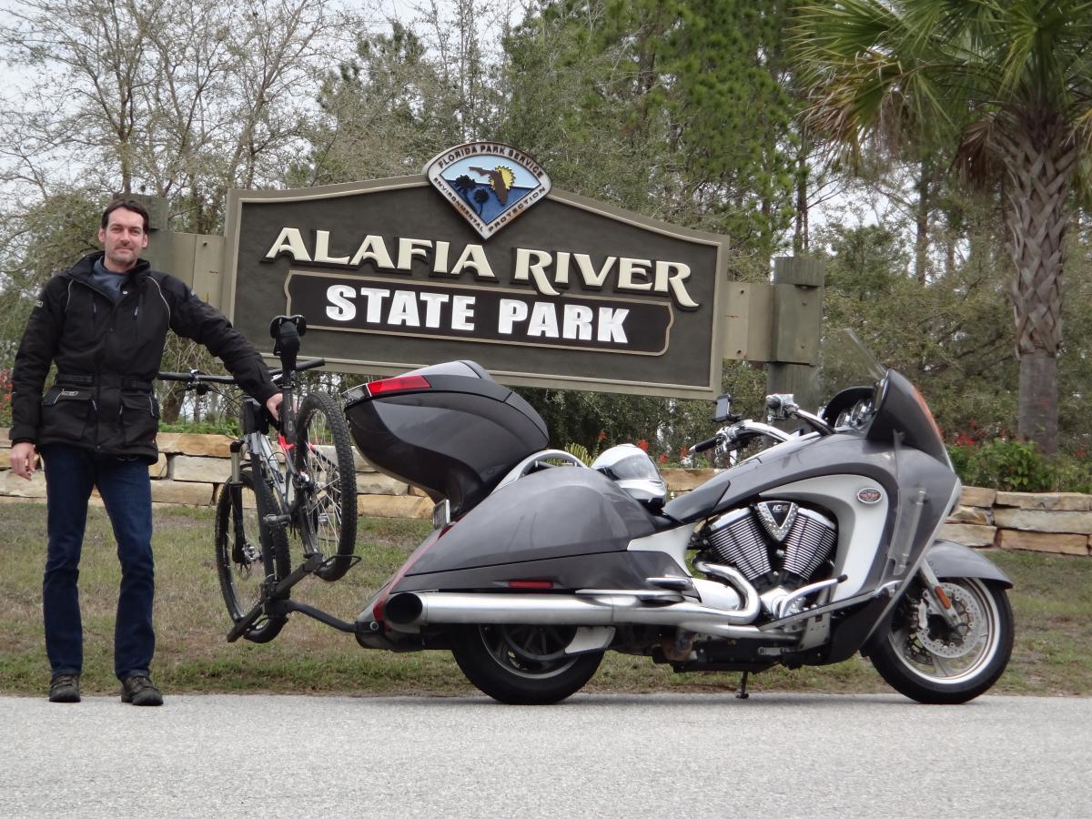 A man standing next to a motorcycle with a bike attached to the back, in front of the Alafia River State Park sign. The scene is set in a natural environment with trees in the background, and the man is wearing a dark jacket and jeans. Alafia River State Park mountain bike trail.