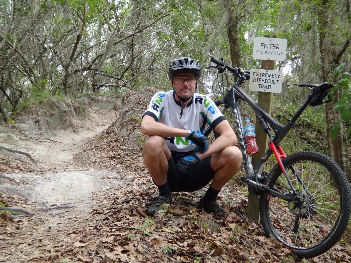 A cyclist in a helmet and athletic gear crouches beside a mountain bike on a dirt trail surrounded by trees. A sign reads "Enter One Way Only" and "Extremely Difficult Trail." The ground is covered with fallen leaves, and there are visible trail markings indicating the rugged terrain. Alafia River State Park mountain bike trail.