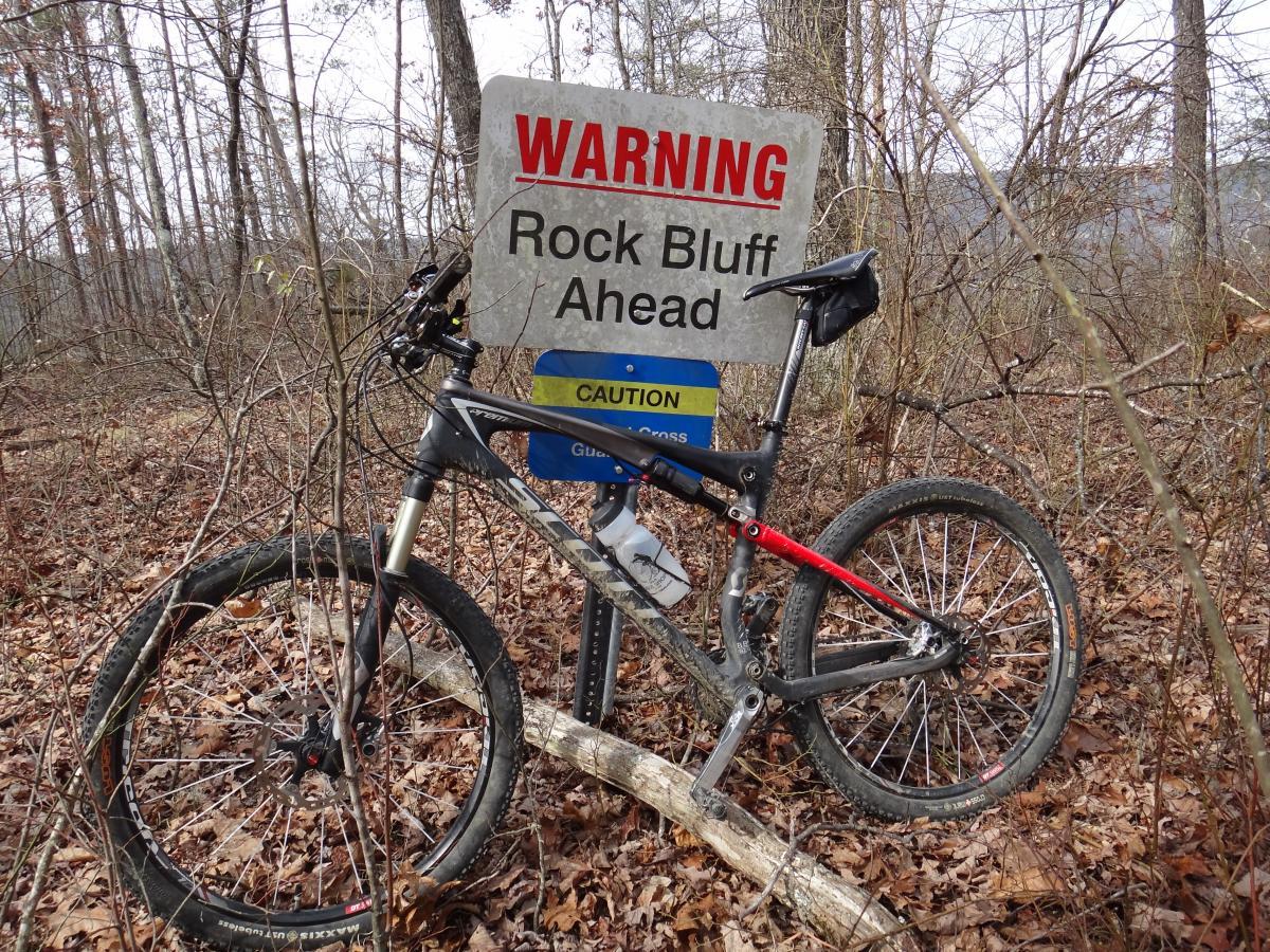 A mountain bike leaning against a caution sign in a forested area, warning of a rock bluff ahead. The surroundings are filled with bare trees and autumn leaves on the ground, indicating a late fall or winter setting. Raccoon Mountain Trail Network mountain bike trail.