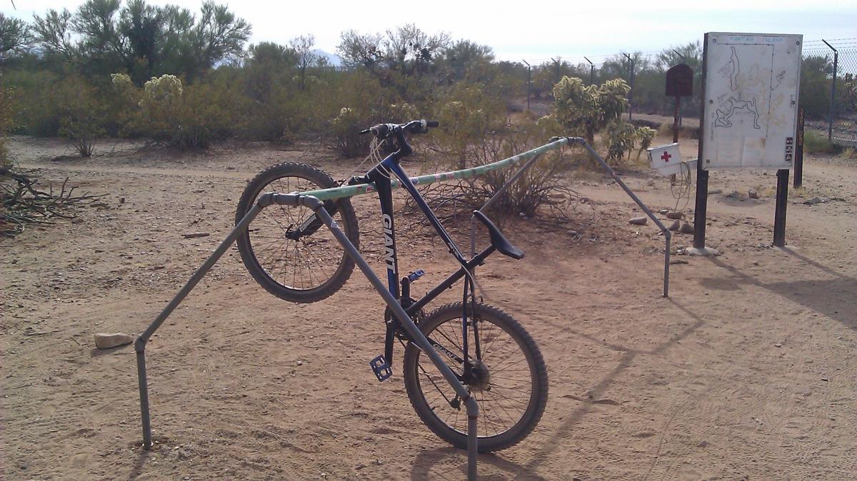 Giant Revel 2: A blue mountain bike is balanced on a makeshift rack made of metal pipes in a desert environment. In the background, there is a trail map sign and sparse vegetation typical of a dry landscape.