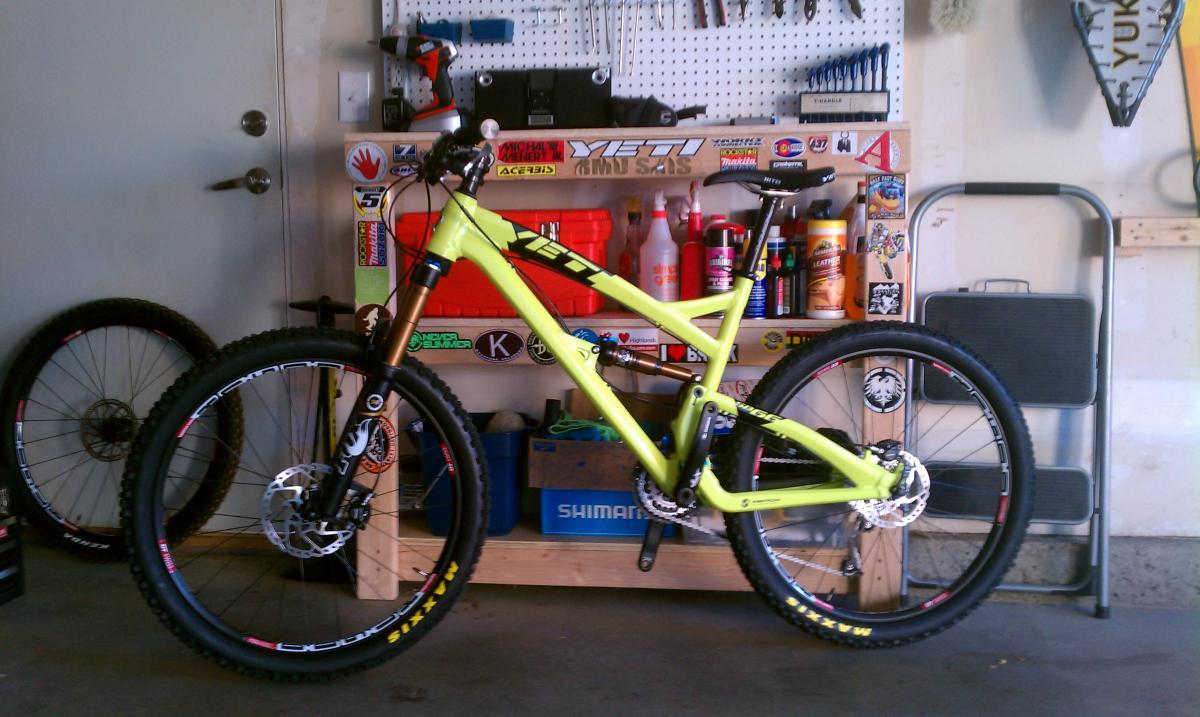 Yeti SB66: A bright yellow mountain bike stands against a wall in a garage, with various tools and supplies in the background. The bike features a sturdy frame and equipped with wide tires, showcasing its suitability for off-road trails. Behind it, there are a variety of colorful bottles and a pegboard filled with tools, while a pair of bike wheels leans against the wall.