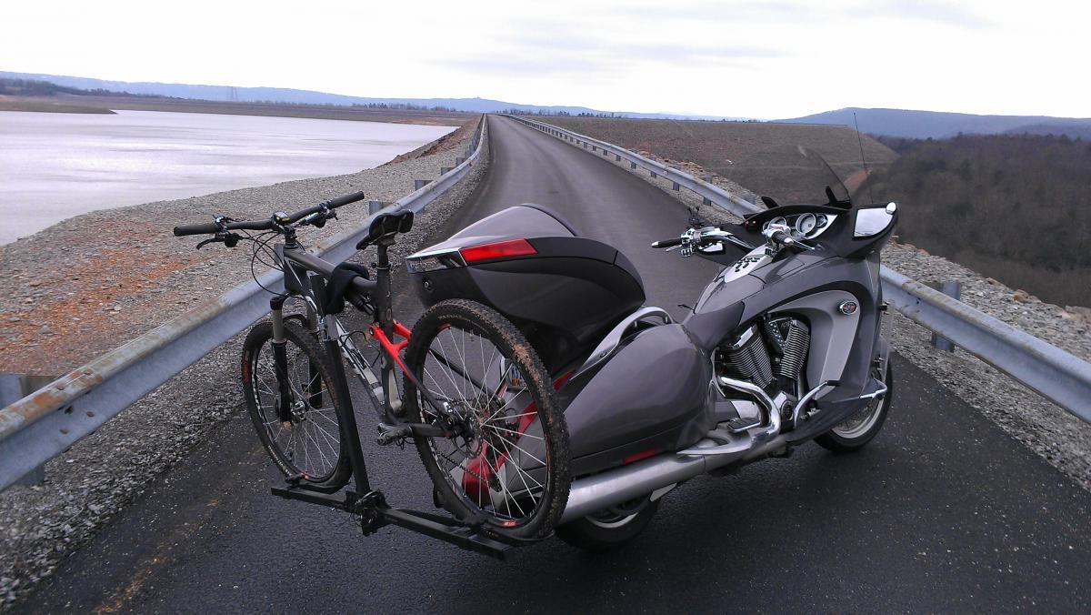 A gray motorcycle with a bike rack attached, holding two bicycles, parked on a road beside a large body of water. The scene features a cloudy sky and hills in the background. Raccoon Mountain Trail Network mountain bike trail.