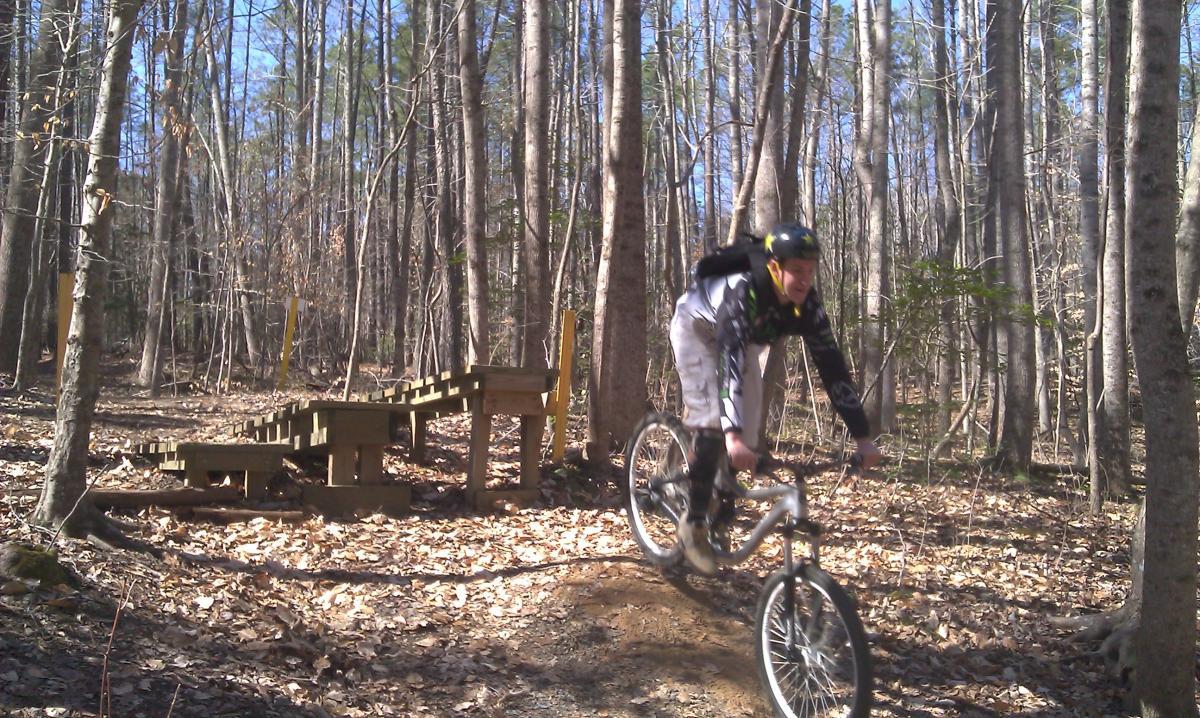 A mountain biker in mid-air, riding on a dirt trail through a forest, with wooden ramps in the background. The scene captures the excitement of biking outdoors on a sunny day, surrounded by tall trees and fallen leaves. Freedom Park (aka:district Park) mountain bike trail.