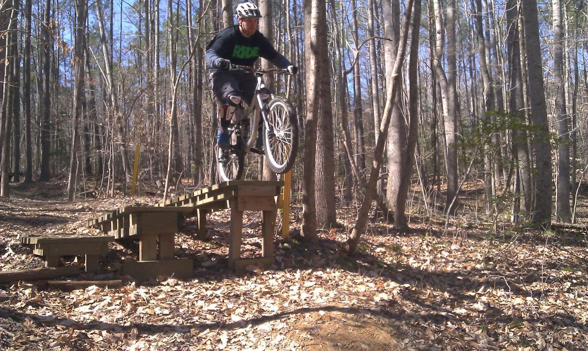 A mountain biker in protective gear performs a jump off a wooden ramp in a forested area, with trees and fallen leaves in the background, on a sunny day. Freedom Park (aka:district Park) mountain bike trail.